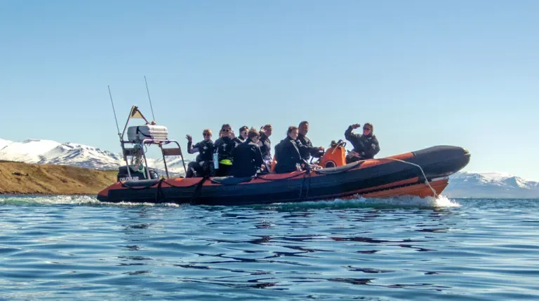 U N E students in wetsuits sit on a small boat with snow-covered mountains in the background